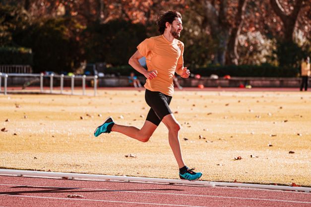 Muž běží po atletickém tartanovém oválu. Na sobě má oranžové triko a černé běžecké kraťasy. Kolem svítí odpolední slunce. 